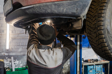 A mechanic in a beanie and work uniform repairing a car's undercarriage on a hydraulic lift in an auto repair shop. Professional vehicle maintenance.