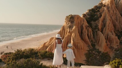 A mother and her son, both dressed in white, walk away from the camera along a coastal path. They are walking towards a view of the ocean with golden cliffs and beaches. - Powered by Adobe