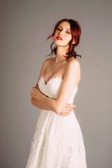 Adorable redhead woman in studio, sensual portrait. Sweetheart neckline double layer bridal white dress. Broidery corset sleeveless evening gown with cut-out. Touching look for an event, elegant style