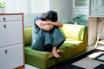 Sad young woman sitting on sofa hugging knees with head down at home