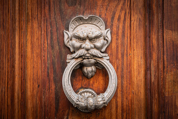 Antique metal door knocker featuring a fierce goblin or demon face biting a large ring. Mounted on...