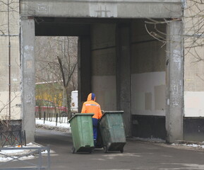 A cleaner in an orange vest moves two mobile green plastic garbage containers in the arch of an apartment building, Podvoysky street, St. Petersburg, Russia, February 24, 2025