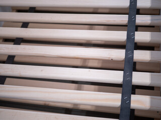 A wooden bed slatted base with evenly spaced light-colored wooden slats held together by black fabric straps. The slats are supported by a bed frame structure.