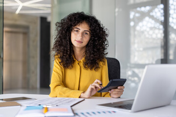 Confident female accountant sits at her desk with documents and laptop, using a calculator to...