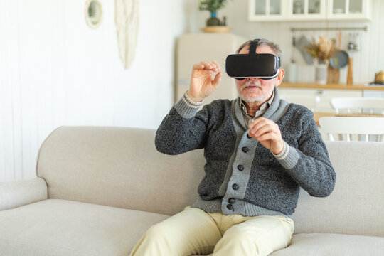 Excited mature senior man wearing using virtual reality metaverse VR glasses headset at home. Grandfather touching air during VR experience on virtual reality helmet. Simulation hi-tech videogame