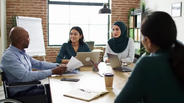 Multiethnic team of diverse business colleagues discussing project during productive meeting in contemporary office, with disabled man in wheelchair explaining document to the team. Teamwork at work