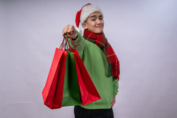 An Asian man wearing a Santa hat, green sweater, and red plaid scarf holds red and green shopping bags with excitement. He smiles joyfully, celebrating holiday shopping against a white background