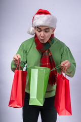 An Asian man wearing a Santa hat, green sweater, and red plaid scarf looks excitedly into holiday shopping bags in red and green. He expresses surprise and joy against a white background.