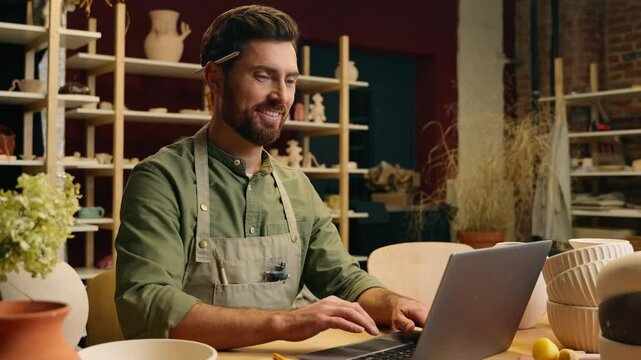 Happy joyful smiling Caucasian man small business owner craftsman businessman potter crock male working laptop typing desktop browsing computer online shopping pottery selling inside workshop studio