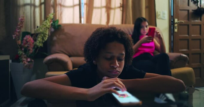 Boy engrossed in smartphone at glass table, girl behind on sofa immersed in phone, illustrating emotional isolation, social disconnection, and digital addiction