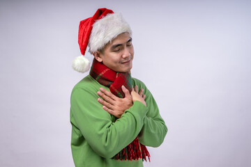 An Asian man wearing a Santa hat, a green sweater, and a red plaid scarf crosses his hands over his chest with a peaceful, He conveys warmth and appreciation against a white background