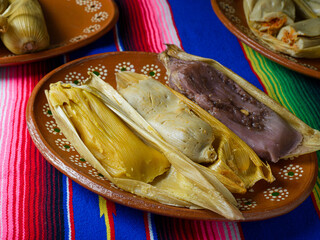 Tamales of different flavors in a clay dish on a colorful serape. Sweet tamales