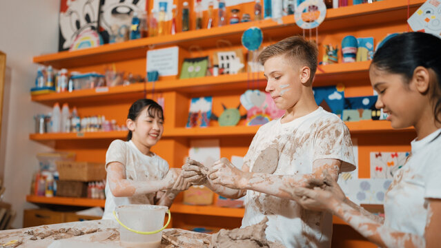 Group of happy diverse children playing clay while wearing muddy shirt at art lesson. Cute highschool girl playing with dough while putting clay on happy boy shirt at pottery workshop. Edification.