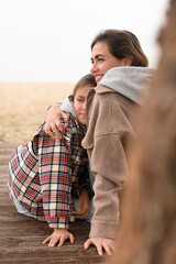 Fototapeta premium A mother and her daughter sit on the sand at the beach, laughing and hugging each other, enjoying their time together despite the chilly weather.
