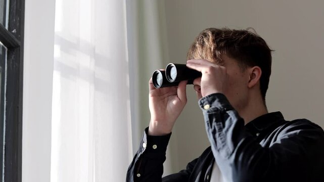 Young man stalker watching through binoculars in front of a window. Man spying or investigating.