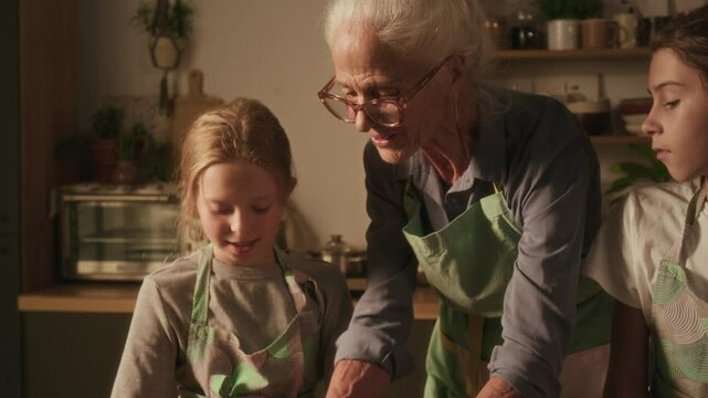 Tilt up view of positive senior woman helping girl flatten dough, her elder brother watching them when all three cooking together