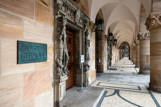 Aufschrift &bdquo;Justizgeb&auml;ude&ldquo; und verzierten Steinfiguren. Der &uuml;berdachte Gang f&uuml;hrt entlang des Eingangsbereichs mit verzierten Holzt&uuml;ren und schmiedeeisernen Laternen. 