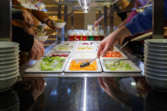 People helping themselves at the colorful salad bar with a healthy selection of fresh vegetables, glasses with colorful desert in the background