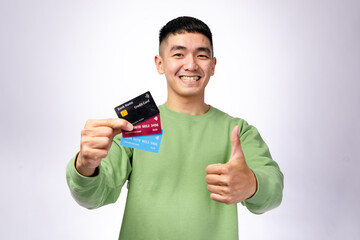 A cheerful Asian man in a green sweatshirt holds three colorful credit cards while giving a thumbs-up. He stands against a plain white background, symbolizing financial success, banking