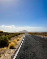 A deserted road stretches into the distance towards a lone mountain under a bright blue sky.