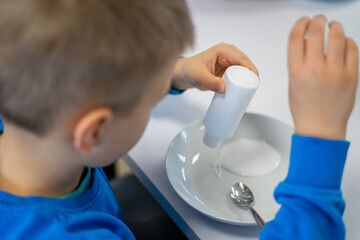A boy in a blue sweater pours white glue into a plate for making slime
