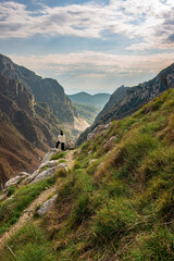 A lone traveler sits on a rocky outcrop, overlooking a vast mountain valley with dramatic cliffs and an open sky. The green slopes contrast with rugged peaks, creating a sense of isolation and wonder.