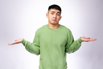 An Asian man in a green sweatshirt shrugs with both hands up and a puzzled expression. He stands against a plain white background, conveying confusion, uncertainty, or a questioning reaction