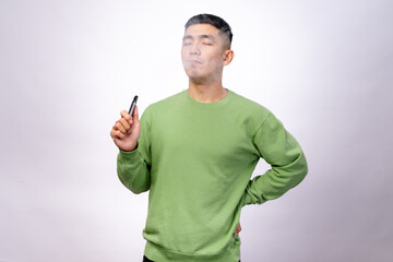 An Asian man wearing a green sweatshirt is vaping, exhaling a visible cloud of vapor with his eyes...