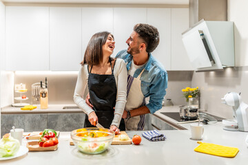 Happy couple preparing vegetable salad, embracing and laughing in modern kitchen