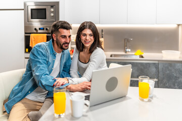 Happy couple browsing internet on laptop in kitchen during breakfast time