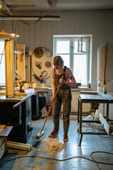 Worker carpenter sweeps sawdust in woodshop after task completion. Handywoman tidying floor with broom, post work cleanup, maintaining cleanliness in workshop, preparing space for upcoming projects