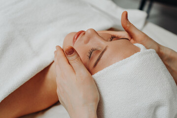 face massage, young attractive caucasian woman, beautiful relaxed lady with closed eyes smiling, white towel, spa therapist doing facial and head massage in beauty salon, skincare and bodycare concept