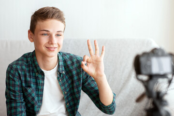 Cute teen guy showing okay while shooting video for his blog, home interior, copy space