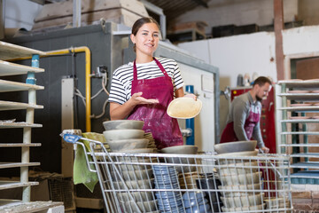 Skillful smiling young female artisan proudly showcasing collection of ceramic plates, highlighting exceptional finish and design in productive pottery workshop