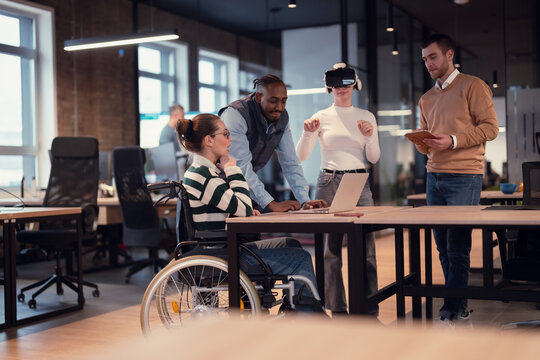 Disabled businesswoman in a wheelchair working in modern coworking office. Team around is using virtual reality goggles, collaborating with the latest technology in an open space startup creative