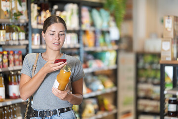 Young woman in supermarket scan QR code bottle of drink. Girl buyer photo information on packaging of bio product.
