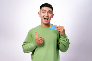 A happy Asian man in a green sweatshirt holds a blue credit card in one hand while giving a...