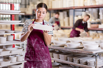 Positive skilled young female artisan standing amidst racks with collection of crafted wares in...