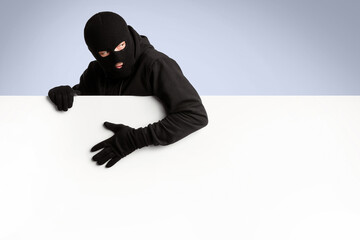 Masked gangster holding white blank advertising panel and looking back, isolated over white studio wall