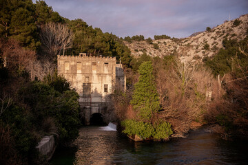 Fototapeta premium An old, abandoned building with a castle-like facade stands by a flowing river, surrounded by lush greenery and rocky hills, bathed in warm sunlight. Solin Croatia