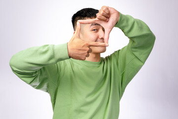 An Asian man in a green sweatshirt makes a rectangle frame with his fingers, looking through it with focus. He stands against a plain white background, symbolizing creativity and photography