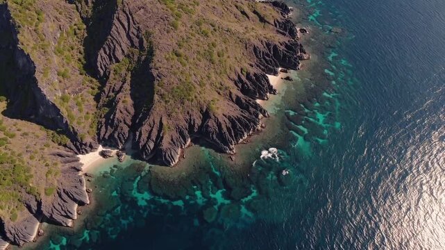 A rugged rocky island is surrounded by deep blue ocean waters, with crystal-clear patches near the coastline revealing submerged reefs and small secluded beaches along the shore