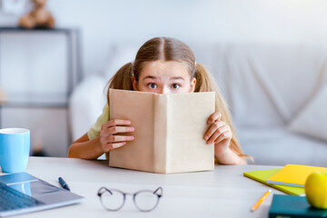 Exam Time And Homework. Portrait of scared girl with wide open eyes hiding her face behind a book,...