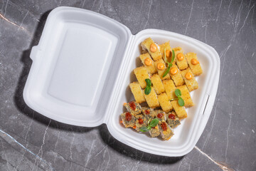 A takeout container with crispy tempura sushi rolls, topped with spicy sauce and fresh green garnish.