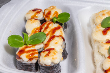 Baked sushi rolls with a golden-brown topping in a styrofoam container, elegantly presented with fresh greens and a drizzle of sauce.