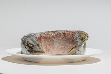 A raw fish with a curved body on a white plate, viewed from the side with strong lighting.