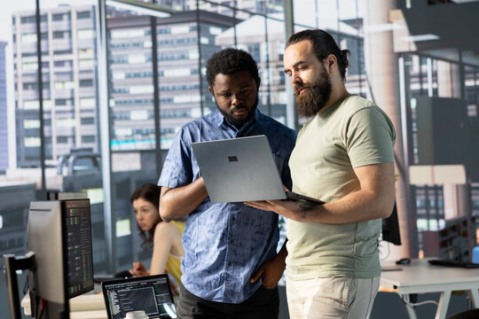 IT specialists in office working together using laptop, testing and deploying programs and systems. Programmer and coworker checking code on notebook, doing brainstorming in workspace