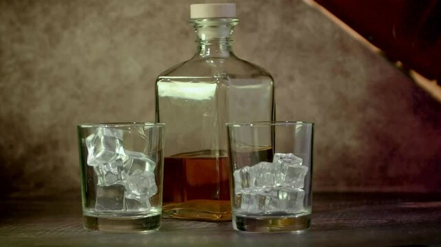 Glasses of whiskey with ice and a bottle of whiskey or brandy on a wooden table. Whiskey is poured into glasses. Antique stylized still life. Slow motion.