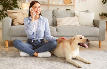 Lifestyle Concept. Portrait of casual smiling woman talking on smartphone, sitting together with cute golden retriever on the floor in living room. Female hipster rubbing her adorable domestic pet