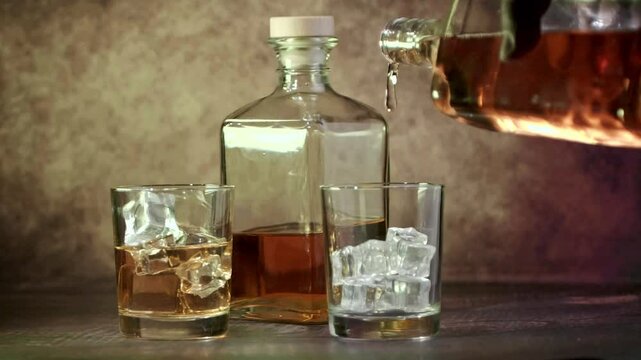 Glasses of whiskey with ice and a bottle of whiskey or brandy on a wooden table. Whiskey is poured into glasses. Antique stylized still life. Slow motion.